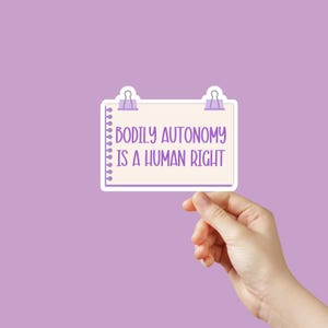 May include: A white paper note with a purple border and two purple paper clips at the top. The note says "Bodily Autonomy is a Human Right". A hand is holding the note.