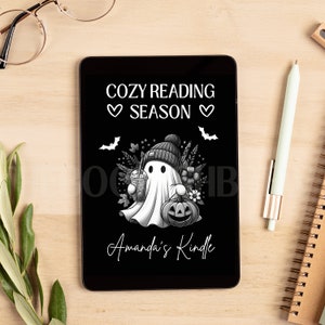 May include: A black and white digital tablet with a ghost holding a pumpkin and a cup of coffee. The text on the screen reads "Cozy Reading Season" and "Amanda's Kindle".