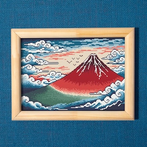 Puede incluir: Un bordado enmarcado que representa el monte Fuji con una cima roja, sobre un cielo de nubes y pájaros. La obra de arte está enmarcada en un marco de madera de color claro, sobre una superficie azul texturizada.