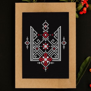 Pode incluir: Um padrão de ponto cruz do tridente ucraniano, um símbolo tradicional da Ucrânia, sobre um fundo de tecido preto. O tridente é bordado em fio branco e vermelho. O tecido é emoldurado com uma moldura de cartão marrom.