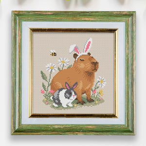 May include: A cross-stitch embroidery framed in green wood, depicting a capybara wearing bunny ears and a black and white rabbit sitting next to it. The capybara and rabbit are surrounded by flowers and a bee.