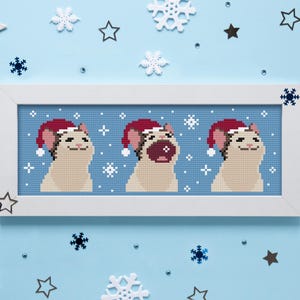 May include: A framed cross-stitch featuring three cats wearing Santa hats. The cats are light beige with red and white hats, set against a blue background with snowflakes and stars. The frame is white.