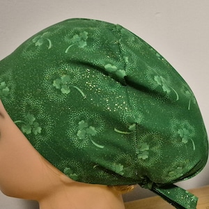 May include: Green fabric head covering with a shamrock pattern. The head covering is tied at the back and is displayed on a mannequin head. The fabric has a repeating pattern of shamrocks and small white dots.