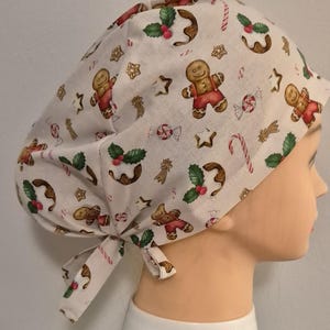May include: A white scrub hat with a Christmas gingerbread man and candy cane print. The hat has a tie at the back.