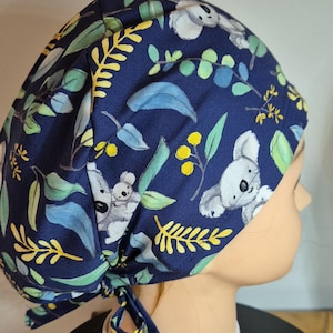 May include: A navy blue scrub cap featuring a pattern of gray koalas, green leaves, and yellow floral accents. The cap is tied at the back and is designed to cover the hair. The fabric has a watercolor-like appearance.