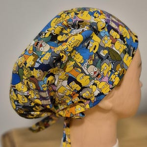 Puede incluir: Un gorro quirúrgico estampado en amarillo, azul y gris con personajes de dibujos animados. El gorro está atado en la parte trasera con lazos de tela a juego. El diseño incluye varios personajes.