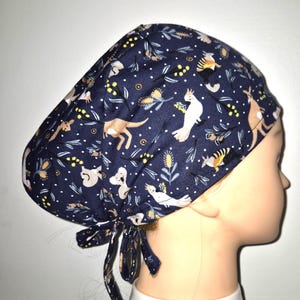 May include: A blue scrub hat with a white background featuring a repeating pattern of Australian animals, including koalas, kangaroos, and cockatoos. The hat has a tie at the back.