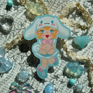 May include: A sticker featuring a cartoon character with orange hair, blue eyes, and a white hat with long ears. The character is wearing a light blue dress and holding a pink cupcake. Surrounding the sticker are various light blue and clear decorative elements.