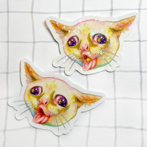 May include: Two stickers of a cat with a surprised expression and its tongue sticking out. The cat's eyes are wide and have glitter stars inside. The cat's fur is yellow and white.