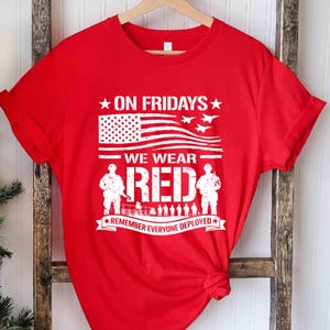 Może przedstawiać: Czerwona koszulka z białą grafiką. Grafika zawiera tekst "ON FRIDAYS WE WEAR RED" z amerykańską flagą, myśliwcami i sylwetkami żołnierzy. Tekst "REMEMBER EVERYONE DEPLOYED" jest również zawarty.