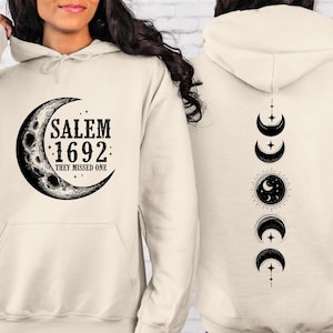 Könnte beinhalten: Cremefarbener Kapuzenpullover mit einem Mond-Grafik-Motiv und dem Text "SALEM 1692: THEY MISSED ONE" auf der Vorderseite. Auf der Rückseite sind Mondphasen-Illustrationen abgebildet. Er hat eine Vordertasche und eine Kapuze mit Kordelzug.