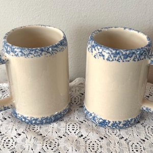 May include: Two cream-colored ceramic mugs with blue floral trim on the rim and handle. The mugs have a cylindrical shape and a sturdy handle. The blue floral pattern adds a decorative touch to the mugs, suitable for beverages.