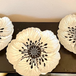 May include: Three white, flower-shaped decorative plates with black centers and accents. The plates are arranged on a dark surface, showcasing their intricate petal designs. The floral theme adds a touch of elegance.