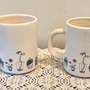 May include: Two white ceramic mugs with a hand-drawn plant design. Each mug features a row of potted plants in various colors, including red, green, and blue. The mugs have a curved handle and a smooth, glossy finish.