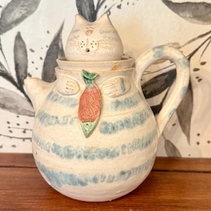 May include: A ceramic teapot with a cat-shaped lid. The teapot is white with blue stripes and a fish-shaped accent. The handle and spout are also blue. The cat lid has a pink nose and whiskers.