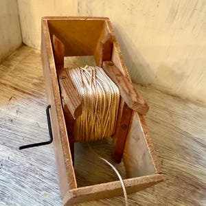 May include: A vintage wooden twine dispenser with a spool of beige twine. The dispenser has a rectangular box shape with a handle and wooden supports. The twine is visible, and a length of twine extends from the dispenser.