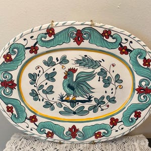 May include: Oval ceramic platter with a hand-painted rooster design. The white platter features a teal border with red and teal floral accents. A yellow border surrounds the central rooster image, which is teal with a red comb.