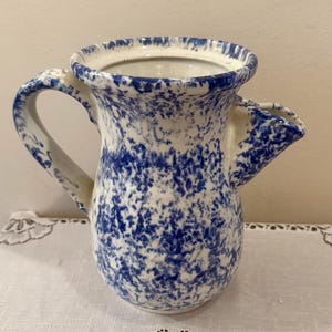 May include: A ceramic pitcher featuring a white base with a blue sponge-painted or speckled pattern. The vessel has a wide, flared rim, a curved handle on one side, and a distinct pouring spout on the opposite side, creating a functional design.