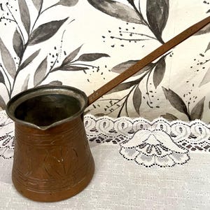 May include: A copper coffee pot with a long handle. The pot has an ornate design and is sitting on a white lace tablecloth.