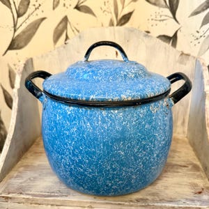 May include: A vintage blue enamelware pot with a lid and black handles. The pot has a speckled white pattern and is sitting on a distressed wooden shelf. The pot is a classic kitchenware item.