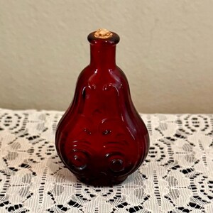May include: A small, ruby red glass bottle with a cork stopper. The bottle has a bulbous body with embossed swirl and floral designs. The bottle's shape is reminiscent of a pear. The cork stopper is a light brown color.