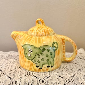 May include: A ceramic teapot with a lid, handle, and spout. The teapot is yellow with orange vertical stripes. A green pig with black hooves and polka dots is painted on the side. The teapot is sitting on a white lace tablecloth.