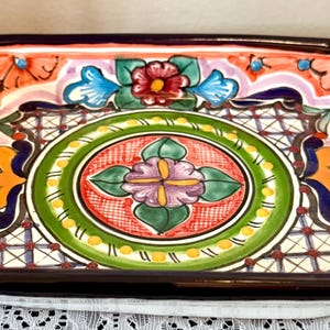 May include: A square ceramic dish featuring a hand-painted design with a central floral motif in purple, green, and red, surrounded by orange and blue patterns. The rim is finished with a thick, dark blue border, creating a vibrant, colorful look.