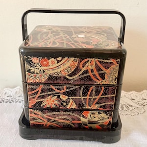 May include: A three-tiered black lacquer bento box with a handle, featuring a decorative pattern of red, gold, and white circular motifs and intertwined ribbons across the exterior surfaces of each stackable container and the base.