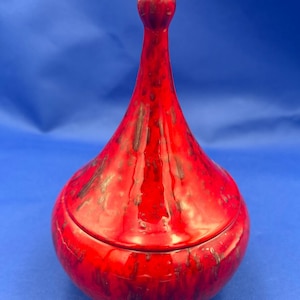 May include: A red ceramic jar with a lid. The jar has a bulbous base and a tall, slender neck that tapers to a rounded top. The red glaze has dark brown streaks. The jar is likely used for storage or decoration.