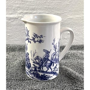 May include: White ceramic pitcher with a blue pastoral scene. The pitcher features a handle and a spout for easy pouring. The blue design depicts figures on horseback, trees, and other elements of a rural landscape. A classic and decorative piece.