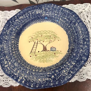 May include: A blue and white ceramic bowl with a speckled glaze. The bowl features a hand-painted design of an apple tree with a ladder and a basket of apples.