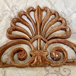 May include: A decorative wall ornament featuring a symmetrical, fan-shaped crest with curved, scroll-like extensions on both sides. The piece has a textured, weathered brown finish with visible signs of wear and paint distress throughout.