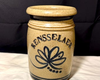 May include: Tan stoneware jar with a dark blue floral design and the word "RENSSELAER" in a curved font. The jar has a dark blue band around the base and rim. The jar is cylindrical with a slightly flared rim.
