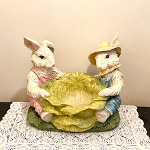 May include: A decorative Easter figurine featuring two white rabbits holding a large, yellow-green cabbage. One rabbit wears pink overalls, the other wears blue overalls and a yellow hat. The base is green.