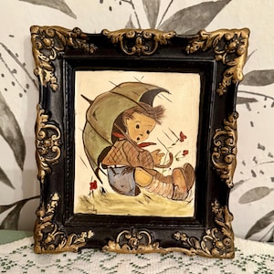 May include: A framed painting depicts a child under an umbrella in the rain. The artwork features a black frame with gold accents. The child is wearing a brown jacket, blue pants, and brown boots. The painting has a vintage aesthetic.