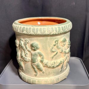 May include: A cylindrical, light green planter with a textured surface and a terracotta interior. The planter features a relief design of cherubs holding a garland around the circumference. The rim and base have decorative molding.