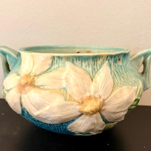 May include: A ceramic bowl featuring a turquoise blue background with a textured surface and raised white floral relief patterns. The bowl has two side handles and a wide opening, with yellow centers on the white flowers and a smooth interior.