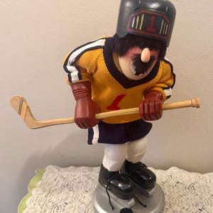 May include: A decorative hockey player figurine wearing a yellow jersey with white and navy blue accents, a black helmet with the number 1, and black skates. The figurine is holding a hockey stick and stands on a silver base.