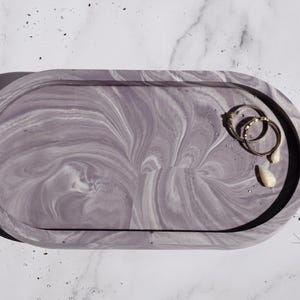 Könnte beinhalten: Ein ovales Tablett aus Beton mit einem Marmormuster in Grautönen und Weiß. Das Tablett hat eine glatte, polierte Oberfläche und ist perfekt für die Aufbewahrung von Schmuck, Schlüsseln oder anderen kleinen Gegenständen.