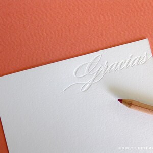 May include: A white card with the word "Gracias" embossed in a cursive script. The card is on a coral background.