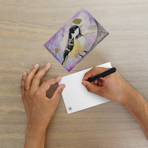 Può includere: Una stampa artistica di uccelli con un uccello nero, bianco e giallo su uno sfondo viola. Una persona sta scrivendo su una carta bianca vuota con una penna nera. La scena è ambientata su una superficie di legno marrone chiaro.
