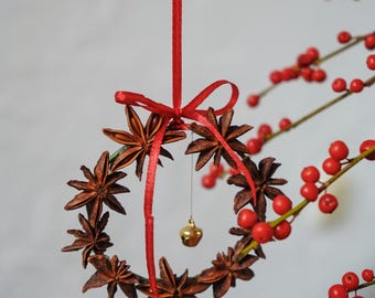 May include: A red ribbon hangs from a branch of red berries, holding a star anise wreath with a small gold bell hanging from the bottom.