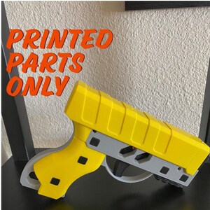 May include: A 3D printed yellow and gray toy gun. The gun is printed in two parts and has a black trigger. The text "PRINTED PARTS ONLY" is visible in the image.