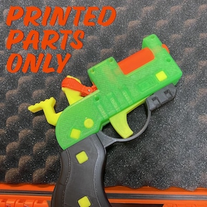 May include: A 3D printed toy gun in green, black, and orange. The gun has a yellow trigger and a black handle. The text "PRINTED PARTS ONLY" is visible in the background.