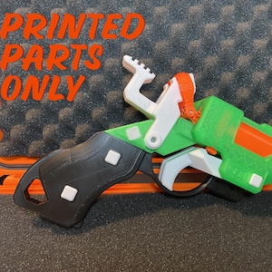 May include: A 3D printed toy gun with a green, white, and orange color scheme. The gun is in a foam-lined case. The text "PRINTED PARTS ONLY" is visible in the image.