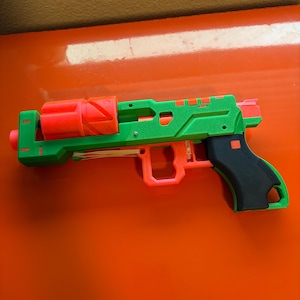 May include: A green and orange toy gun with a black handle. The gun has a spring-loaded mechanism and is designed for recreational use.