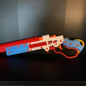 May include: A red, white, and blue toy gun with a yellow and blue handle. The gun has a textured surface and a gear-like mechanism.