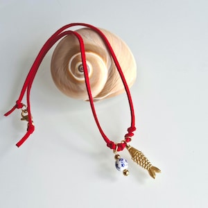 May include: A red cord necklace featuring a gold-toned fish pendant and a small round bead with a blue and white floral pattern. The necklace includes a gold-toned lobster clasp closure and is displayed against a large, spiraled tan seashell.