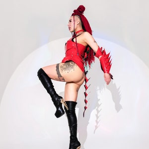 May include: A woman wearing a red leather outfit with a long, spiky tail. She is wearing black thigh-high boots and has red hair with a braid. She is posing with one leg raised and her arm extended.