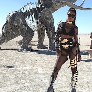 May include: A woman wearing a black and gold outfit with a metal harness and fishnet stockings. She is standing in front of a large metal sculpture of an elephant.